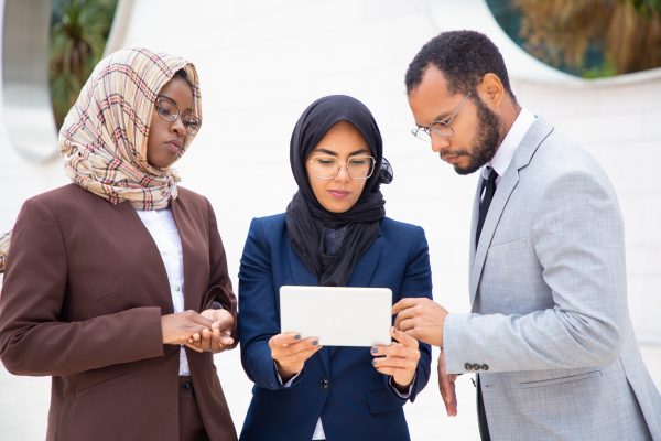Serious business team watching project presentation on tablet outside. Businessman and Muslim businesswomen standing outdoors and using tablet together. Diverse team concept